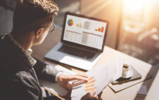 Young finance market analyst in eyeglasses working at sunny office on laptop while sitting at wooden table.Businessman analyze document in his hands.Graphs and diagramm on notebook screen.Blurred